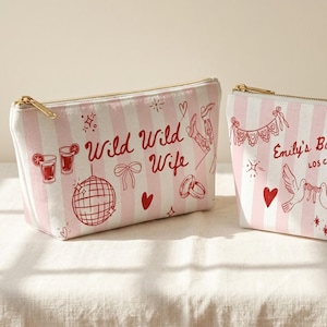 May include: Two rectangular cosmetic bags with pink and white stripes. One bag has the text "Wild Wild Wife" and the other says "Emily's Bachelorette Los Cabos 2026". Both bags have gold zippers and red illustrations.