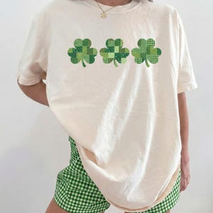 Puede incluir: Camiseta color crema con tres diseños de tréboles verdes. Los tréboles tienen un patrón de patchwork. Combinada con shorts de cuadros verdes y blancos, ideal para el Día de San Patricio o cualquier ocasión informal.
