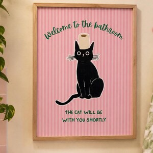Op de afbeelding: Een ingelijste wandkunstprint met een zwarte kat met een wc-rol op zijn kop, tegen een roze gestreepte achtergrond. De tekst luidt "Welcome to the bathroom" en "THE CAT WILL BE WITH YOU SHORTLY" in groene letters.