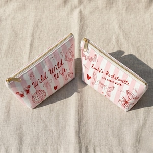 May include: Two pink and white striped cosmetic bags with gold zippers. One bag reads "Wild Wild Wife" with red illustrations. The other bag reads "Emily's Bachelorette Los Cabos 2026" with red illustrations.