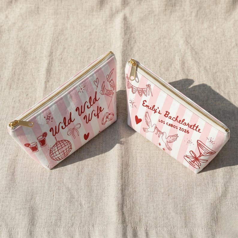 May include: Two pink and white striped cosmetic bags with gold zippers. One bag reads "Wild Wild Wife" with red illustrations. The other bag reads "Emily's Bachelorette Los Cabos 2026" with red illustrations.