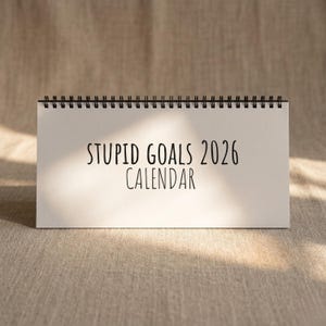 以下が含まれることがあります： 黒いスパイラル綴じの白い卓上カレンダー。テキスト「STUPID GOALS 2026 CALENDAR」は黒で印刷され、表紙の中央に配置されています。カレンダーは、ニュートラルな質感の背景に配置されています。