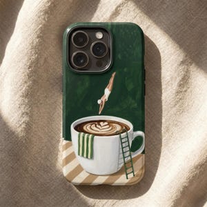 May include: Green phone case featuring a whimsical illustration of a diver plunging into a coffee cup. The cup has a latte art design and a green and white striped towel. A small ladder is placed on the side of the cup.