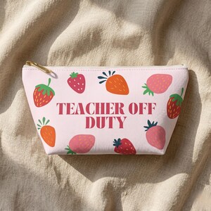 Puede incluir: Un estuche con cremallera de color rosa claro con un patrón repetitivo de fresas rojas y naranjas con hojas verdes. El texto "TEACHER OFF DUTY" está impreso en letras mayúsculas de color rosa oscuro en el centro de la parte frontal.