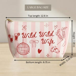 May include: A large, pink and white striped cosmetic bag with the text "Wild Wild Wife" in red. The bag features illustrations of cocktails, a disco ball, a bow, a heart, cowboy boots, and wedding rings. Top length is 12.6 inches.