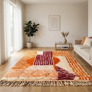 May include: A large, rectangular rug with a geometric pattern in orange, white, and burgundy. The rug has a shaggy texture and a fringe along the bottom edge. The rug is in a living room with a sofa and a potted plant.