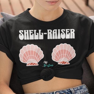 Może przedstawiać: Czarny t-shirt z napisem "SHELL-RAISER" białymi literami. Dwie różowe muszle znajdują się pod napisem.