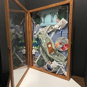 May include: A wooden shadow box with a glass front, displaying baby items. Inside are a gray onesie with a bear graphic, a baby hat, footprints, and other keepsakes. The box is open, revealing a reflection.