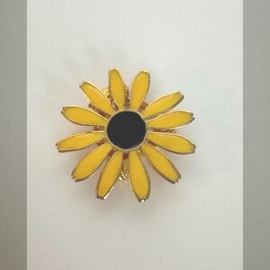 May include: A gold-toned metal daisy pin with yellow petals and a black center. The pin is a flower shape, with a bright, cheerful design. The pin is likely a fashion accessory.