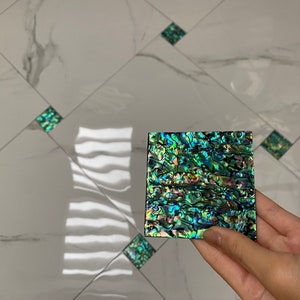 May include: A close-up of a square iridescent green and blue tile with a mother-of-pearl finish. The tile is being held in a hand.