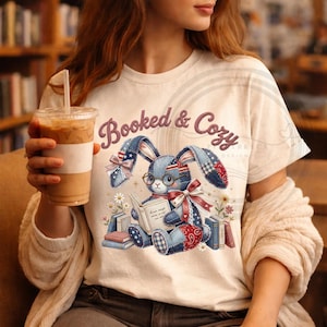 May include: Cream t-shirt featuring a denim bunny reading a book, wearing glasses and a bow tie. The text "Booked & Cozy" is arched above the bunny. The bunny is surrounded by books and flowers.