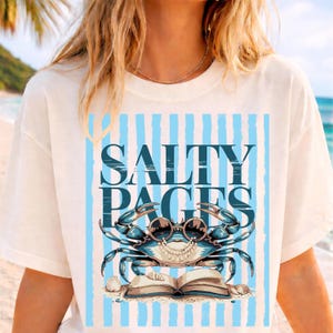 May include: Cream t-shirt with a blue crab wearing sunglasses and reading a book. The text "SALTY PAGES" is above the crab, with blue vertical stripes in the background. A seashell is at the bottom of the design.