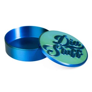Op de afbeelding: Een ronde, metallic blauwe container met een bijpassend deksel. Het deksel heeft een lichtgroen, iriserend oppervlak met de in reliëf aangebrachte tekst "Dia Stuff" in een donkerblauw cursief lettertype. De container heeft een gladde opening.