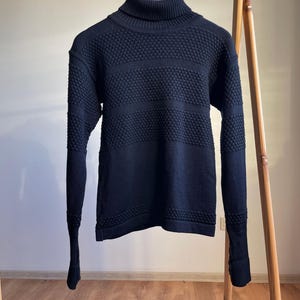 Puede incluir: Un suéter negro de cuello alto con detalles de punto texturizado. El suéter presenta un cuello alto, puños y dobladillo acanalados, con patrones horizontales y texturizados en el pecho y las mangas. La prenda está colgada en una percha de madera.