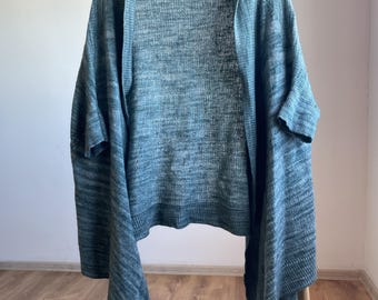 May include: A teal-colored open cardigan with short sleeves hangs on a wooden hanger. The knit fabric has a subtle texture and a label that reads "FREEDOM OF STYLE". The garment is displayed against a light-colored wall and wooden floor.
