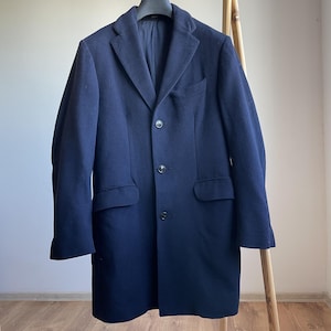 May include: A navy blue overcoat with a classic design, featuring a notched lapel, three buttons, and two front pockets. The coat is hanging on a wooden clothing rack. The coat's length extends to mid-thigh.