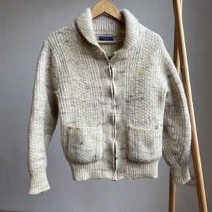 Könnte beinhalten: Cremefarbene Strickjacke mit Reißverschluss und zwei Vordertaschen. Der Pullover hat einen Schalkragen und hängt an einem hölzernen Kleiderständer. Das Strickware hat ein strukturiertes Aussehen mit dunkleren Farbtupfern.