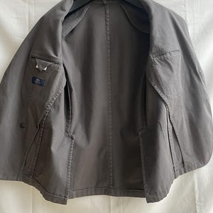 May include: A gray blazer with a classic design, featuring two front pockets and an interior pocket. The jacket is open, revealing the interior lining and construction details. The blazer is hanging on a black hanger.