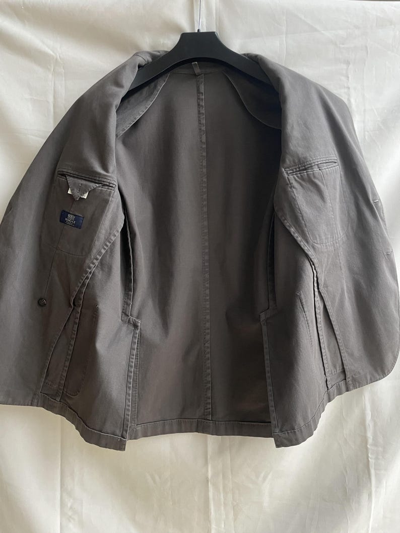 May include: A gray blazer with a classic design, featuring two front pockets and an interior pocket. The jacket is open, revealing the interior lining and construction details. The blazer is hanging on a black hanger.