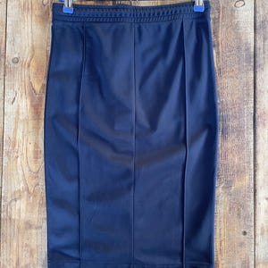 May include: A dark navy blue pencil skirt featuring a smooth, athletic-style fabric with a ribbed elastic waistband and two vertical stitched seams running down the front. The skirt is suspended from a metal hanger with two blue plastic clips.