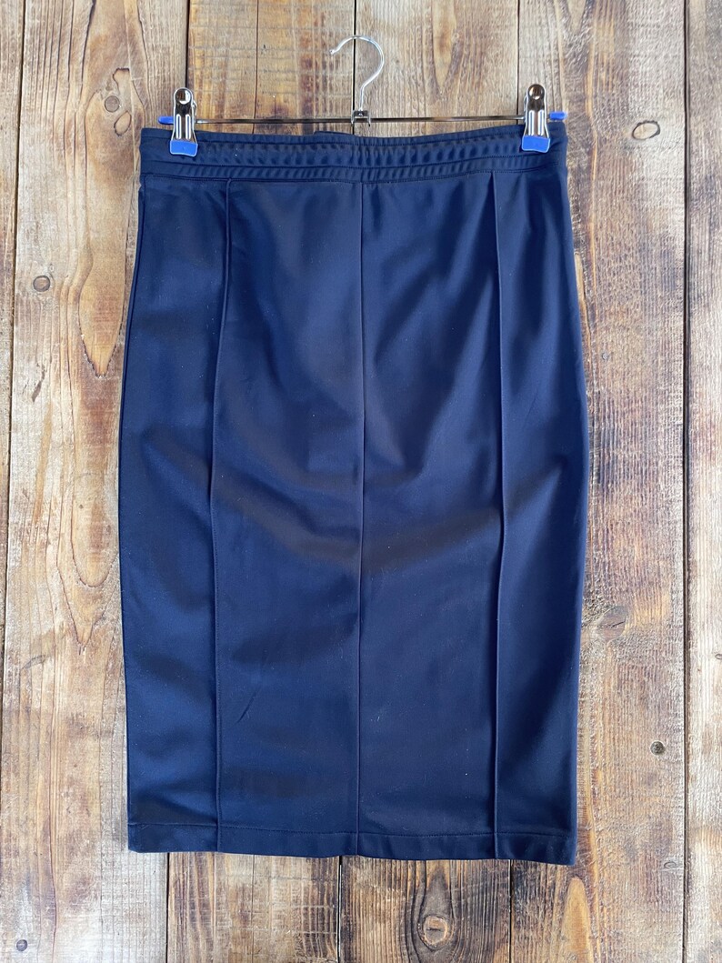 May include: A dark navy blue pencil skirt featuring a smooth, athletic-style fabric with a ribbed elastic waistband and two vertical stitched seams running down the front. The skirt is suspended from a metal hanger with two blue plastic clips.