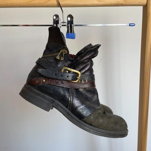 May include: Black leather ankle boot with a distressed appearance, featuring a buckle and strap detail. The boot has a zipper closure and a contrasting olive green suede toe. It hangs from a silver hanger on a wooden rack.