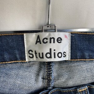 Puede incluir: Primer plano de unos vaqueros azules con el nombre de la marca "Acne Studios" impreso en una etiqueta blanca. La etiqueta est&aacute; cosida a los vaqueros. Los vaqueros cuelgan de una percha met&aacute;lica.