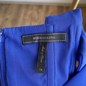 Puede incluir: Primer plano de un vestido azul vibrante. La etiqueta dice "BCBGMAXAZRIA" en texto blanco sobre una etiqueta rectangular negra. Debajo de la etiqueta hay una etiqueta m&aacute;s peque&ntilde;a que dice "Made in China" y el n&uacute;mero "08".