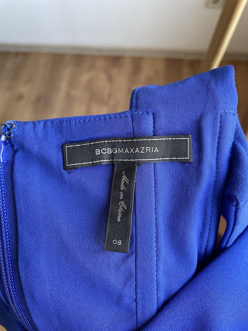 Puede incluir: Primer plano de un vestido azul vibrante. La etiqueta dice "BCBGMAXAZRIA" en texto blanco sobre una etiqueta rectangular negra. Debajo de la etiqueta hay una etiqueta m&aacute;s peque&ntilde;a que dice "Made in China" y el n&uacute;mero "08".