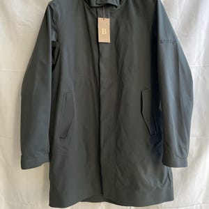 May include: A dark green, hooded coat with a button closure and two front pockets. The coat has long sleeves and a tag with the letter "B" hanging from the collar. The word "B TECH" is printed on the sleeve.