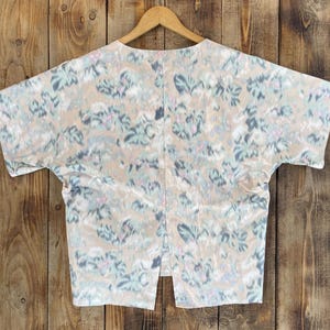 Puede incluir: Una blusa de manga corta con base beige y un patr&oacute;n abstracto de pinceladas en tonos azul, verde y rosa apagados. La prenda presenta una silueta amplia y cuadrada con una abertura vertical en el centro del dobladillo inferior, sobre una percha.
