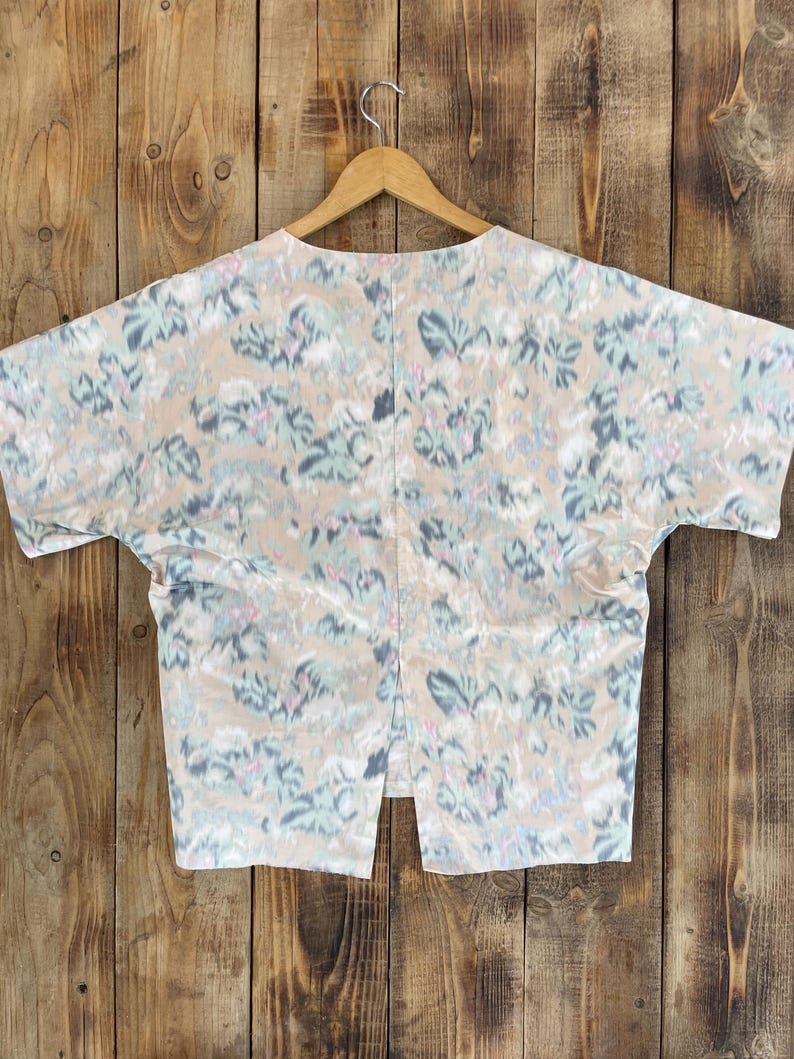 Puede incluir: Una blusa de manga corta con base beige y un patr&oacute;n abstracto de pinceladas en tonos azul, verde y rosa apagados. La prenda presenta una silueta amplia y cuadrada con una abertura vertical en el centro del dobladillo inferior, sobre una percha.
