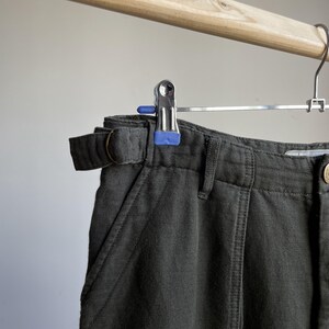 May include: Close-up of dark green pants with a belt and a metal hanger with blue accents. The pants have a belt loop and a pocket. The fabric appears to be a sturdy material.