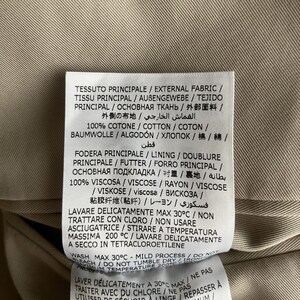 May include: Close-up of a clothing tag with text in multiple languages. The tag is white with black text and is attached to a tan fabric. The tag provides washing instructions and fabric composition.