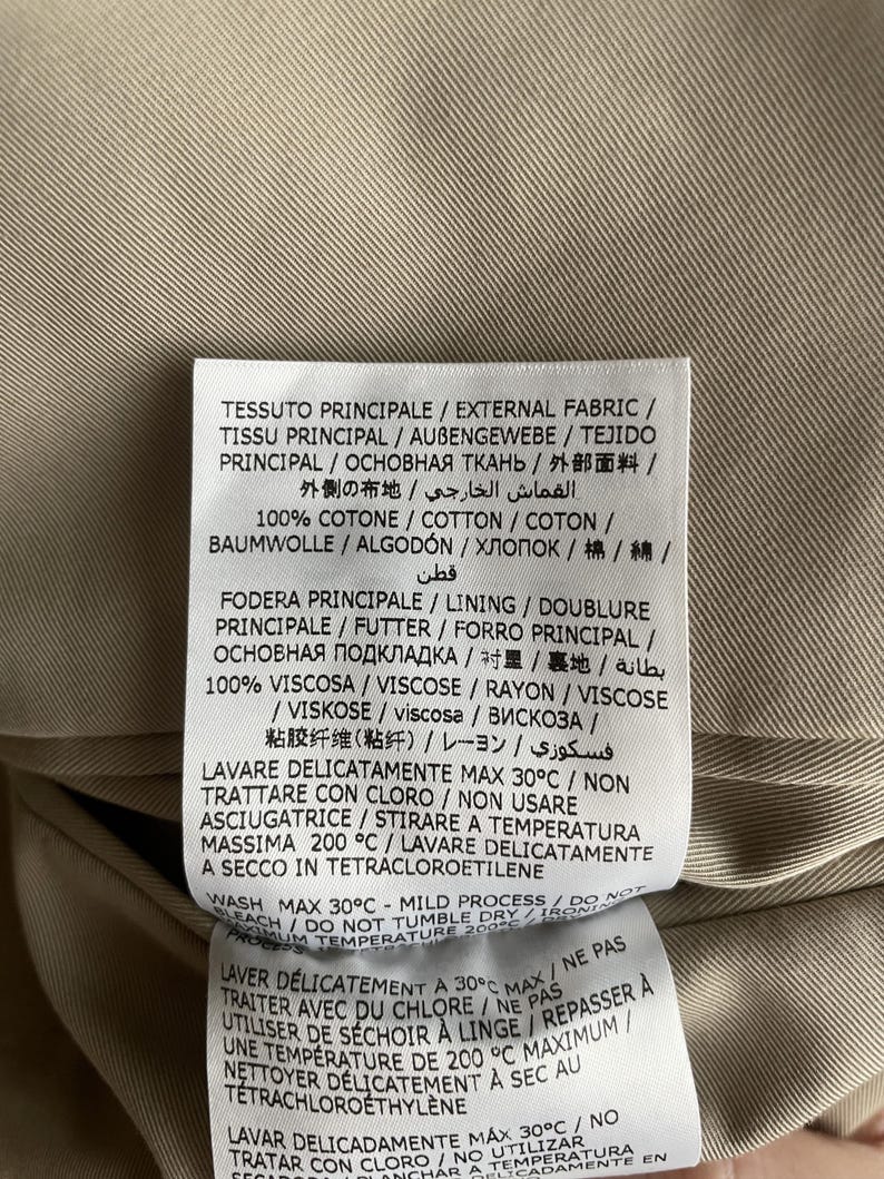 May include: Close-up of a clothing tag with text in multiple languages. The tag is white with black text and is attached to a tan fabric. The tag provides washing instructions and fabric composition.