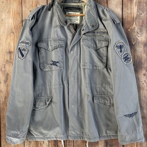 May include: A sage green field jacket featuring a high collar, four front pockets with button flaps, and a front zipper closure. The jacket is adorned with various embroidered patches and text including Alpha Industries, featuring a drawstring hem.