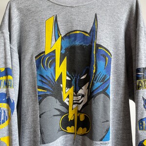 May include: A grey sweatshirt featuring a large graphic print of Batman's face with a lightning bolt behind him. The sleeves have smaller Batman graphics.