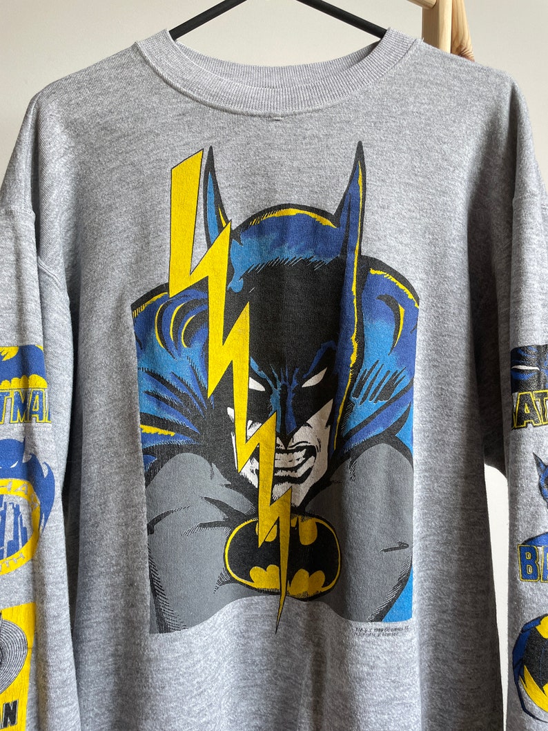 May include: A grey sweatshirt featuring a large graphic print of Batman's face with a lightning bolt behind him. The sleeves have smaller Batman graphics.