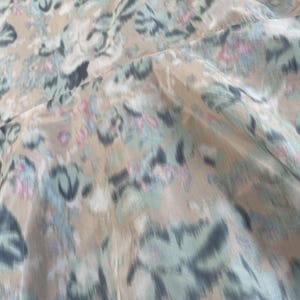 Puede incluir: Una blusa de color beige con un estampado floral difuminado en tonos azul claro, gris oscuro y rosa suave. La prenda tiene un cuello redondo y se muestra en una percha de madera, destacando una tela ligera con una ca&iacute;da fluida.