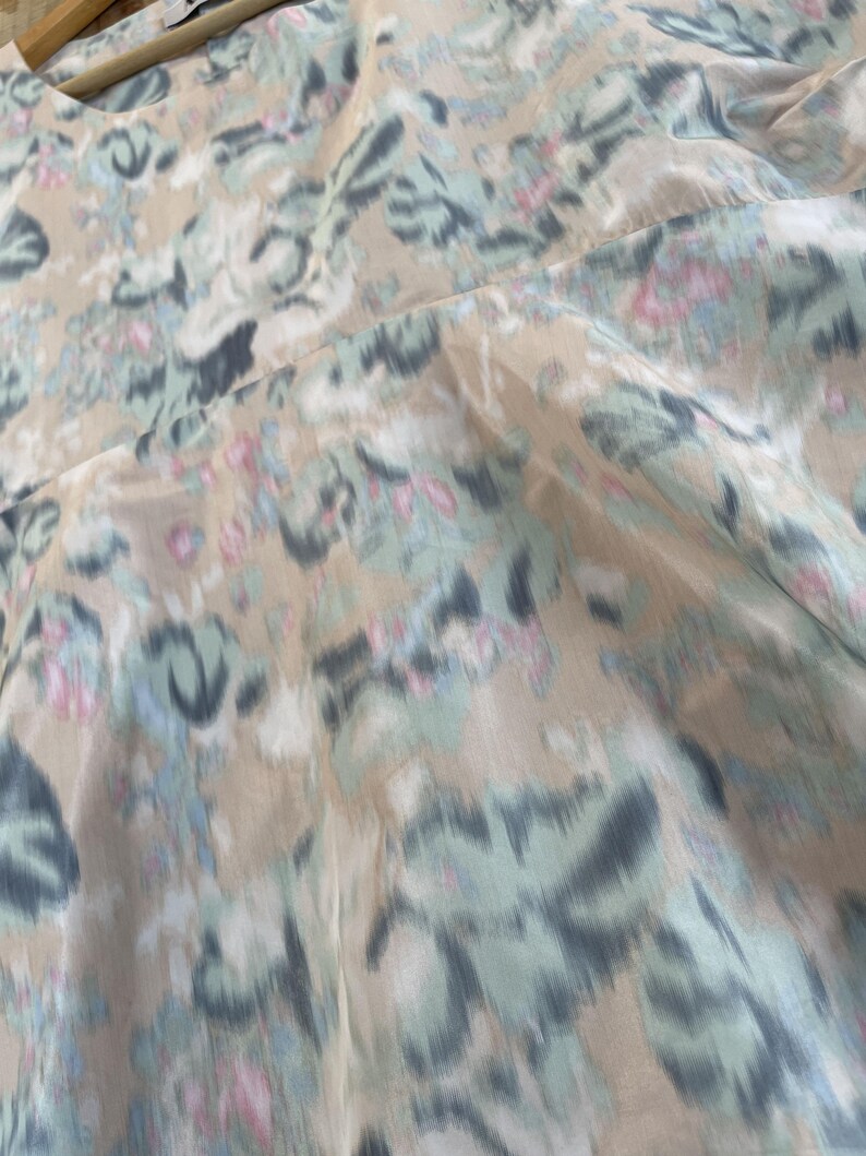 Puede incluir: Una blusa de color beige con un estampado floral difuminado en tonos azul claro, gris oscuro y rosa suave. La prenda tiene un cuello redondo y se muestra en una percha de madera, destacando una tela ligera con una ca&iacute;da fluida.
