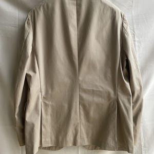 May include: A tan blazer with a notched lapel, hanging from a black hanger. The back of the jacket is visible, showing a tailored fit with a center vent and pleats. The fabric appears to be a lightweight material.