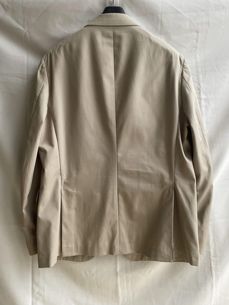 May include: A tan blazer with a notched lapel, hanging from a black hanger. The back of the jacket is visible, showing a tailored fit with a center vent and pleats. The fabric appears to be a lightweight material.