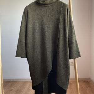 Peut inclure: Tunique en tricot vert olive avec un col roul&eacute; et des manches trois-quarts. La tunique pr&eacute;sente un ourlet asym&eacute;trique et est pr&eacute;sent&eacute;e sur un portant en bois. Le tissu semble &ecirc;tre un tricot textur&eacute;.