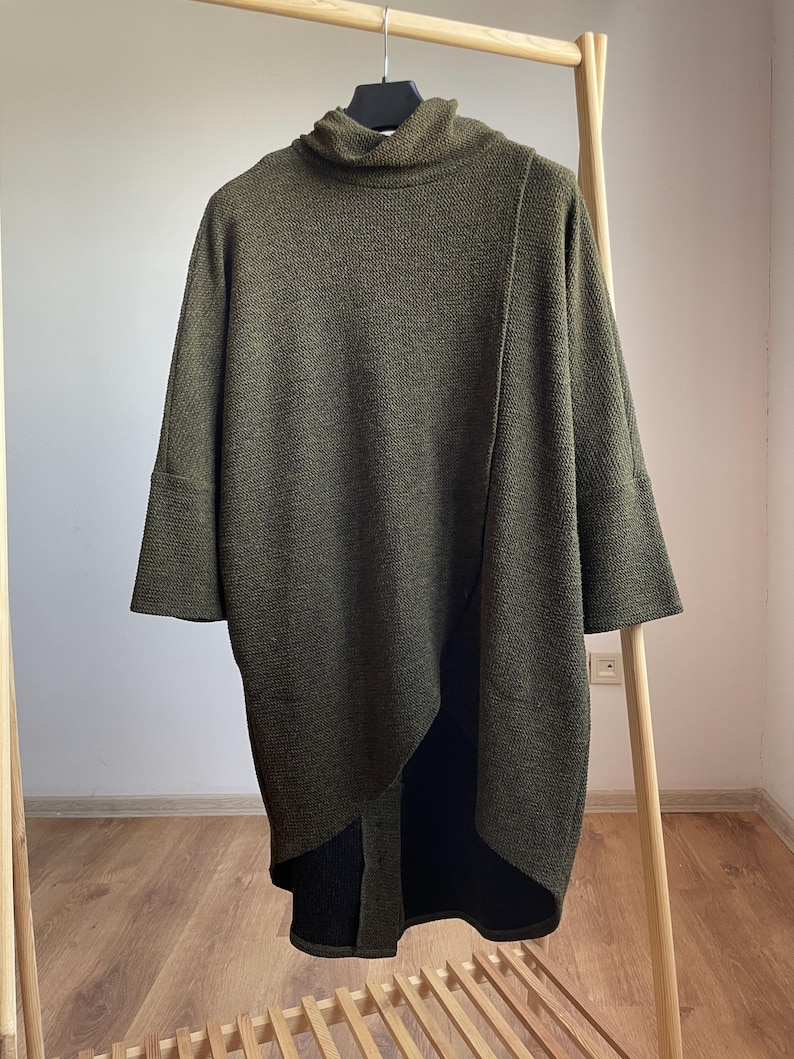 Peut inclure: Tunique en tricot vert olive avec un col roul&eacute; et des manches trois-quarts. La tunique pr&eacute;sente un ourlet asym&eacute;trique et est pr&eacute;sent&eacute;e sur un portant en bois. Le tissu semble &ecirc;tre un tricot textur&eacute;.