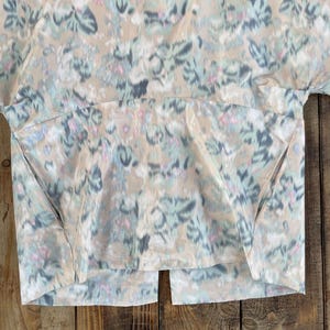 Puede incluir: Una blusa de manga corta con base beige y un patr&oacute;n floral difuminado en tonos azul, gris y rosa. La prenda presenta un cuello redondo, una costura horizontal en el pecho, dos bolsillos laterales y una abertura vertical en el dobladillo.