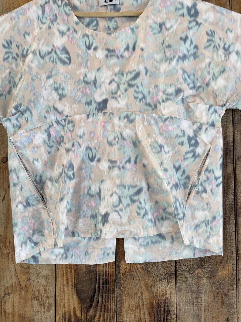 Puede incluir: Una blusa de manga corta con base beige y un patr&oacute;n floral difuminado en tonos azul, gris y rosa. La prenda presenta un cuello redondo, una costura horizontal en el pecho, dos bolsillos laterales y una abertura vertical en el dobladillo.