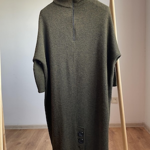Peut inclure: Robe tunique en tricot vert olive avec un col montant et une fermeture &eacute;clair noire. La robe a des manches courtes et des boutons au niveau de l'ourlet. Le v&ecirc;tement est pr&eacute;sent&eacute; sur un portant en bois.