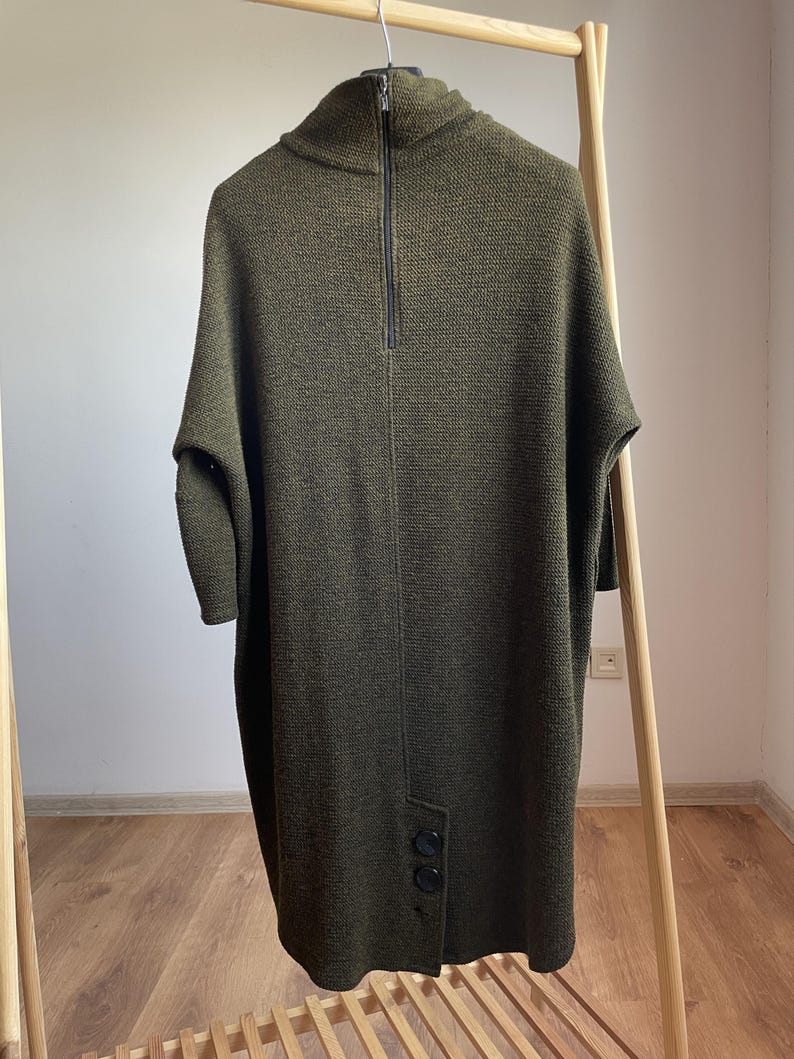 Peut inclure: Robe tunique en tricot vert olive avec un col montant et une fermeture &eacute;clair noire. La robe a des manches courtes et des boutons au niveau de l'ourlet. Le v&ecirc;tement est pr&eacute;sent&eacute; sur un portant en bois.