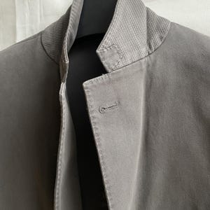 May include: Close-up of a gray jacket with a textured collar and a single button closure. The jacket is made of a smooth fabric and has a tailored fit. The collar is a lighter shade of gray than the body of the jacket.