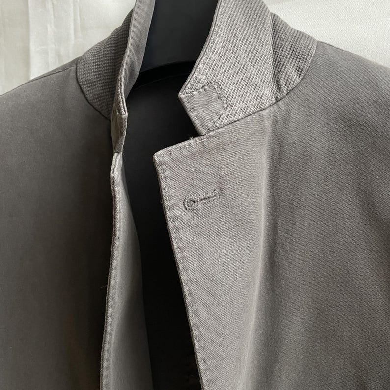 May include: Close-up of a gray jacket with a textured collar and a single button closure. The jacket is made of a smooth fabric and has a tailored fit. The collar is a lighter shade of gray than the body of the jacket.